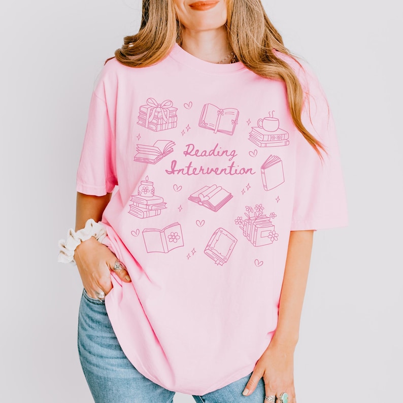 May include: Pink t-shirt featuring a graphic design of books, a cup of coffee, and the text "Reading Intervention".
