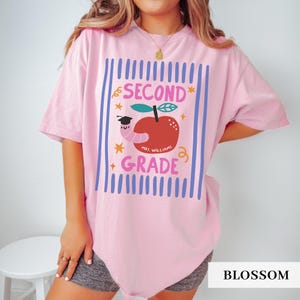 Pu&ograve; includere: T-shirt rosa chiaro con un design di seconda elementare. Il design presenta una mela rossa con un verme con un cappello di laurea, testo e stelle. Il testo dice "SECOND GRADE" e "MRS. WILLIAMS".