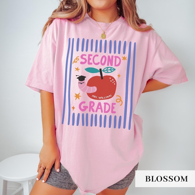 Pu&ograve; includere: T-shirt rosa chiaro con un design di seconda elementare. Il design presenta una mela rossa con un verme con un cappello di laurea, testo e stelle. Il testo dice "SECOND GRADE" e "MRS. WILLIAMS".