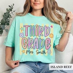 May include: A mint green short-sleeve t-shirt featuring a graphic with vertical yellow stripes. The text reads "THIRD GRADE" in large, colorful, bubble-style letters, with "Mrs. Smith" below. Small star, flower, and smiley face icons surround the text.