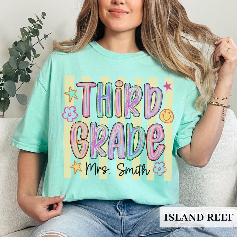 May include: A mint green short-sleeve t-shirt featuring a graphic with vertical yellow stripes. The text reads "THIRD GRADE" in large, colorful, bubble-style letters, with "Mrs. Smith" below. Small star, flower, and smiley face icons surround the text.