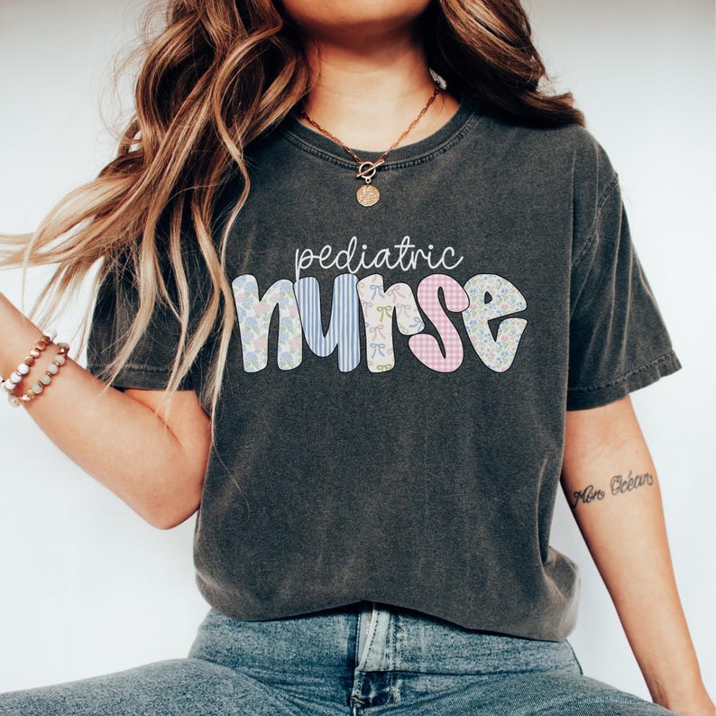 Op de afbeelding: Een donkergrijs T-shirt met de tekst "pediatric nurse" in kleurrijke patchwork letters. De letters zijn versierd met bloemen- en ruitpatronen.