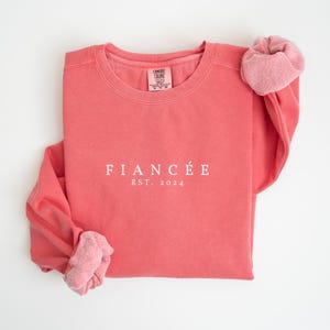 May include: A coral pink sweatshirt with the text "FIANC&Eacute;E EST. 2024" in white.