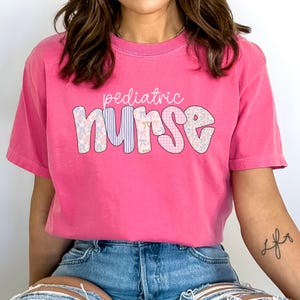 Op de afbeelding: Een roze T-shirt met de tekst "pediatric nurse" in een patchwork-stijl lettertype. De letters zijn versierd met bloemen- en ruitpatronen.