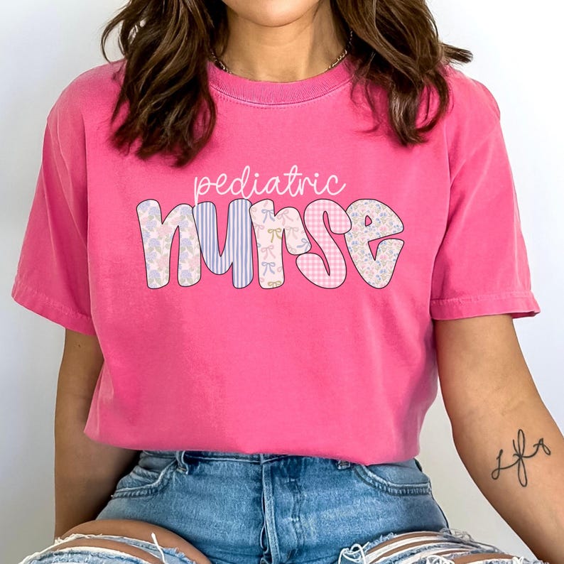 Op de afbeelding: Een roze T-shirt met de tekst "pediatric nurse" in een patchwork-stijl lettertype. De letters zijn versierd met bloemen- en ruitpatronen.