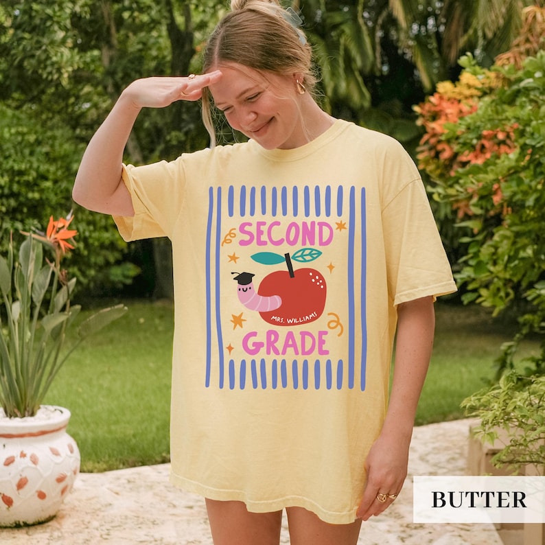 Pu&ograve; includere: T-shirt giallo chiaro con un design grafico per la seconda elementare. Il design include una mela con un verme, le parole "SECOND GRADE" e "MRS. WILLIAM!" in un font colorato e giocoso.