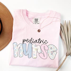 Op de afbeelding: Roze T-shirt met de tekst "pediatric nurse" in een patchwork-stijl met bloemen-, ruit- en streepmotieven.