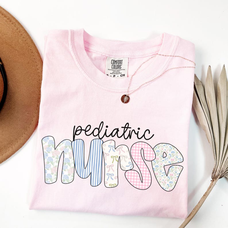 Op de afbeelding: Roze T-shirt met de tekst "pediatric nurse" in een patchwork-stijl met bloemen-, ruit- en streepmotieven.