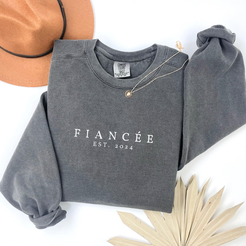 May include: Grey sweatshirt with the text "FIANC&Eacute;E EST. 2024" in white.