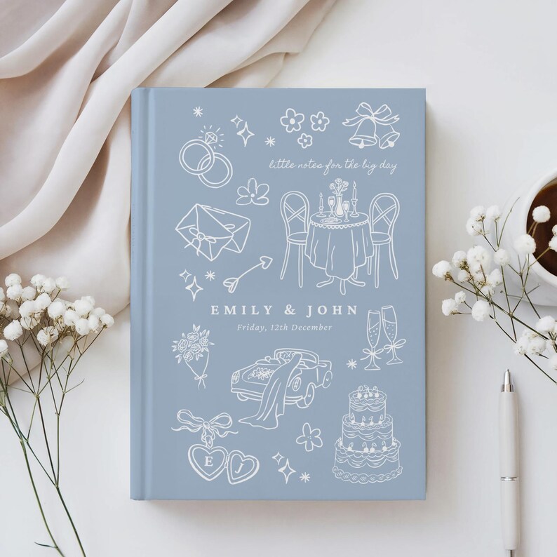 May include: A light blue hardcover journal featuring white line art illustrations of wedding icons including rings, a table setting, a car, and a cake. The text reads "little notes for the big day", "EMILY & JOHN", and "Friday, 12th December".