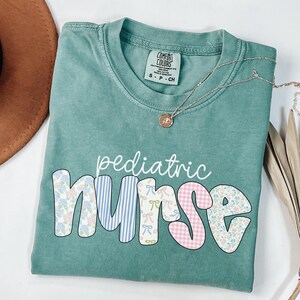 Op de afbeelding: Een tealblauw t-shirt met de woorden "pediatric nurse" in witte letters. Het woord "nurse" is gemaakt van kleurrijke patchwork-vierkanten met bloemen- en ruitpatronen.