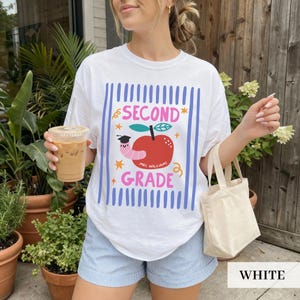 Pu&ograve; includere: T-shirt bianco con un design grafico colorato raffigurante una mela, un verme e le parole "SECOND GRADE". Il design &egrave; incorniciato da strisce verticali blu. La maglietta &egrave; abbinata a pantaloncini a righe blu e bianche.