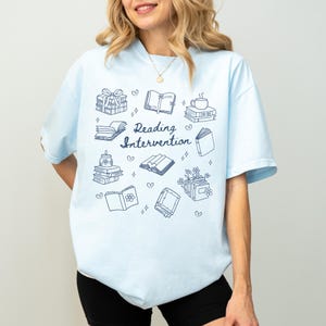 May include: Light blue t-shirt with a graphic design of books, hearts, and the text "Reading Intervention".