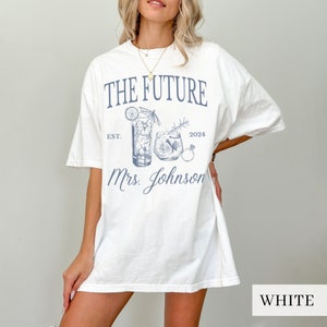 May include: White oversized t-shirt with a blue graphic design featuring two cocktail glasses, a ring, and the text "The Future Est. 2024 Mrs. Johnson".