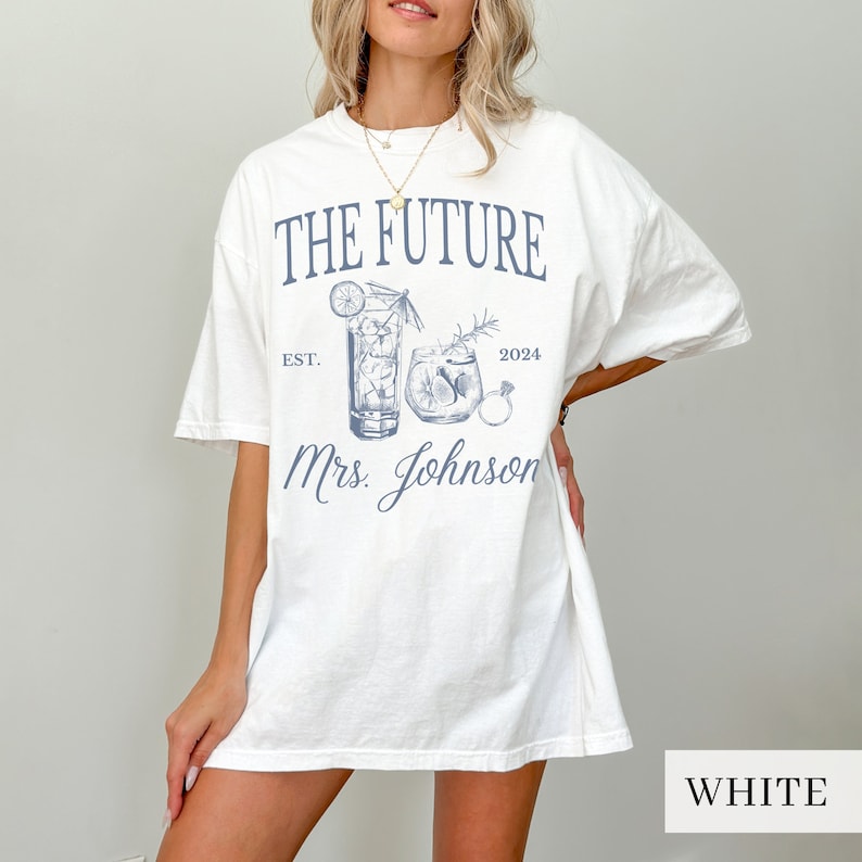 May include: White oversized t-shirt with a blue graphic design featuring two cocktail glasses, a ring, and the text "The Future Est. 2024 Mrs. Johnson".