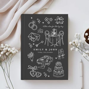 May include: A dark gray hardcover notebook featuring white line art illustrations of wedding icons including rings, a table setting, a car, and a cake. The text reads "little notes for the big day", "EMILY & JOHN", and "Friday, 12th December".