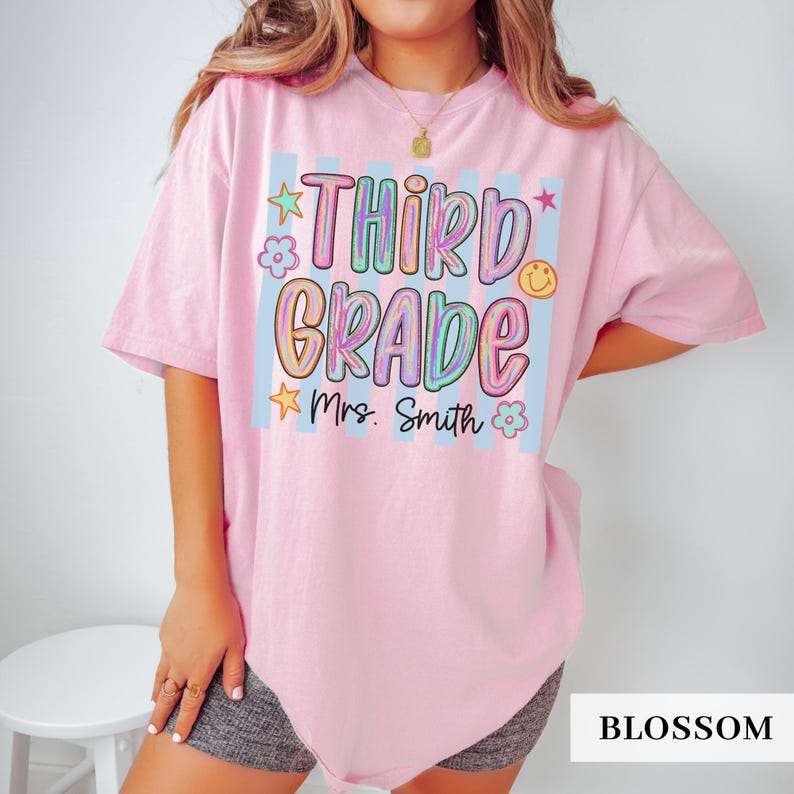 May include: A light pink short-sleeve t-shirt featuring a graphic with vertical light blue stripes and the text "THIRD GRADE" in colorful, bubbly letters. Below the text, "Mrs. Smith" is printed in black script, surrounded by small stars and flowers.