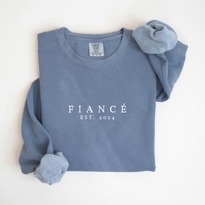 May include: A light blue sweatshirt with the text "FIANC&Eacute; EST. 2024" printed in white on the front.