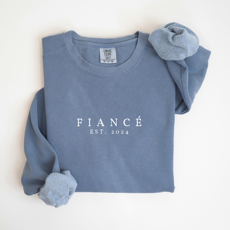 May include: A light blue sweatshirt with the text "FIANC&Eacute; EST. 2024" printed in white on the front.