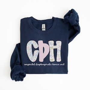 May include: Navy blue sweatshirt with the letters "CDH" in a whimsical font. The "C" is patterned with blue and pink flowers, the "D" with pink gingham, and the "H" with bows. The text "congenital diaphragmatic hernia unit" is below.