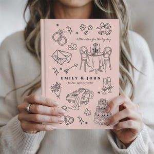 May include: A light pink hardcover journal featuring black line art illustrations of wedding-themed icons including rings, a table setting, a car, a cake, and champagne flutes. The text reads "little notes for the big day", "EMILY & JOHN", and "Friday, 12th December".