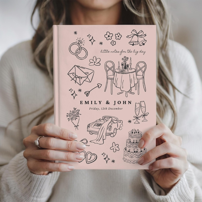 May include: A light pink hardcover journal featuring black line art illustrations of wedding-themed icons including rings, a table setting, a car, a cake, and champagne flutes. The text reads "little notes for the big day", "EMILY & JOHN", and "Friday, 12th December".