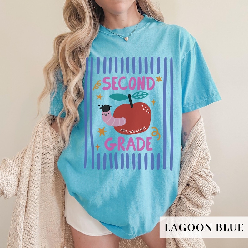 Pu&ograve; includere: T-shirt blu laguna con grafica raffigurante una mela rossa, un verme con cappello di laurea e le scritte "SECOND GRADE" e "MRS. WILLIAM". Il disegno &egrave; incorniciato da linee blu verticali.