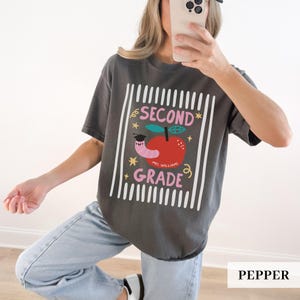 Pu&ograve; includere: T-shirt color pepe con un design grafico per la seconda elementare. Il design presenta una mela rossa con un verme con un cappello di laurea, le parole "SECOND GRADE" e "MR. WILLIAM" in rosa e linee verticali bianche.