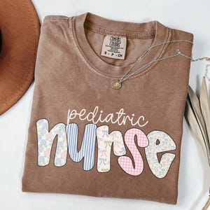 Op de afbeelding: Een bruin T-shirt met de woorden "pediatric nurse" in kleurrijke patchwork letters. De letters zijn gemaakt van verschillende patronen, waaronder bloemen, ruitjes en strepen.