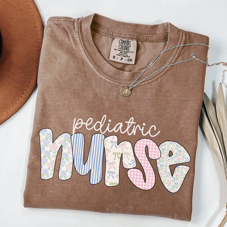 Op de afbeelding: Een bruin T-shirt met de woorden "pediatric nurse" in kleurrijke patchwork letters. De letters zijn gemaakt van verschillende patronen, waaronder bloemen, ruitjes en strepen.