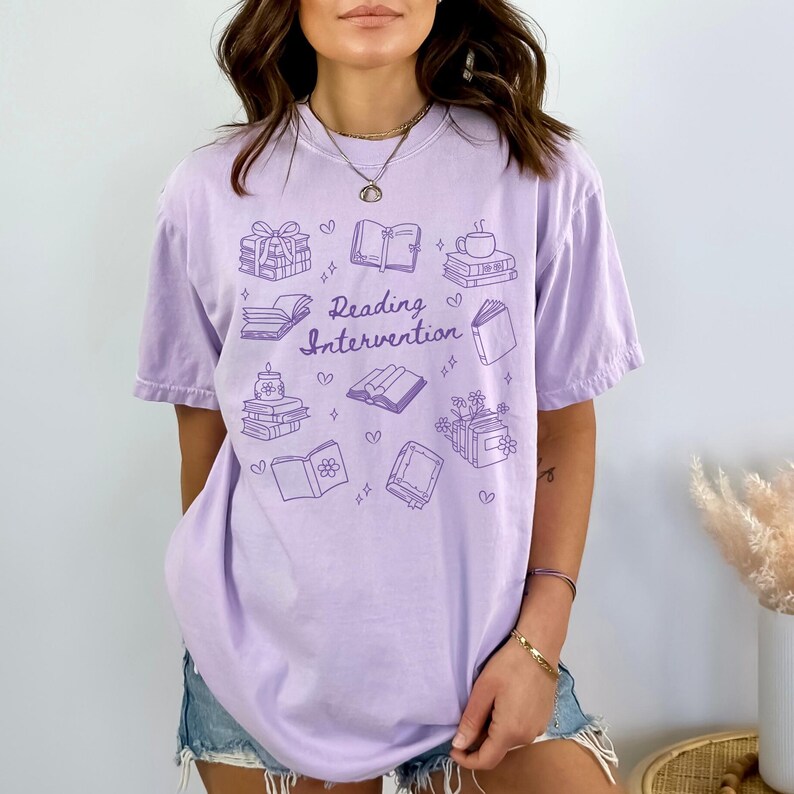 May include: A lavender t-shirt featuring a graphic design of books, hearts, and stars. The text "Reading Intervention" is printed on the shirt.