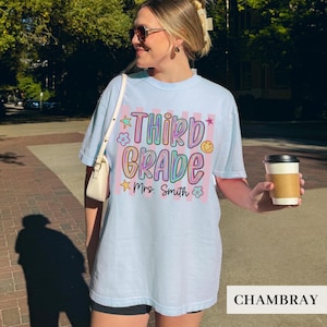 May include: A light blue t-shirt featuring a graphic with vertical pink stripes and the text "THIRD GRADE" in a colorful, bubbly font. Below the text, "Mrs. Smith" is written in black script, surrounded by small star and flower icons.