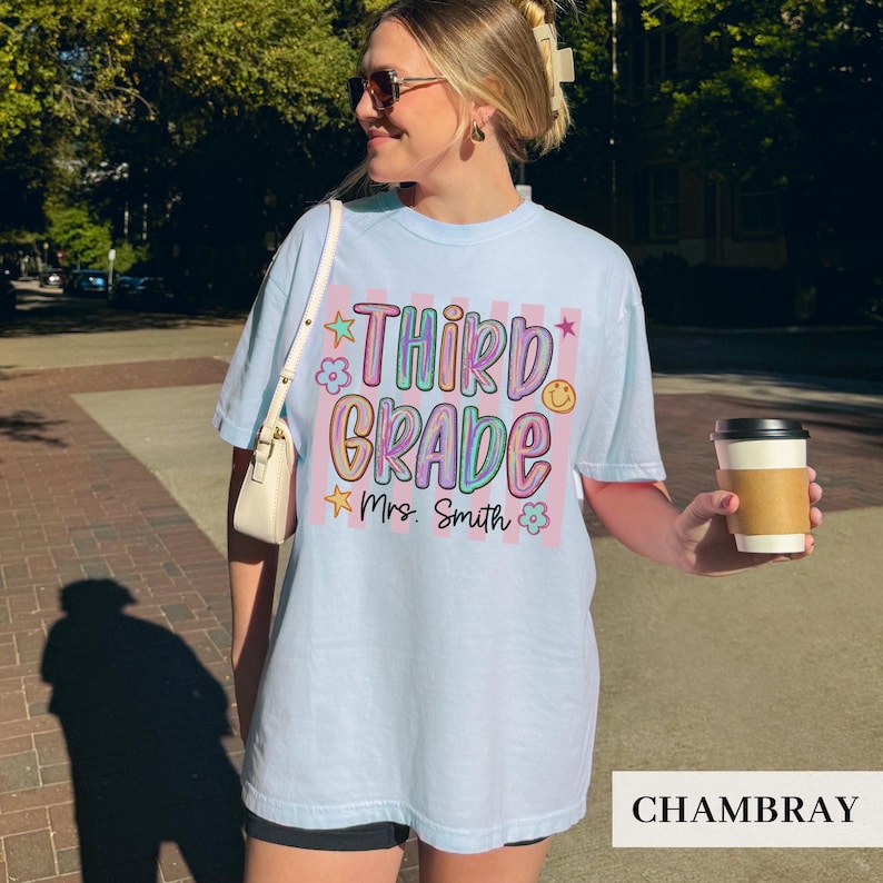 May include: A light blue t-shirt featuring a graphic with vertical pink stripes and the text "THIRD GRADE" in a colorful, bubbly font. Below the text, "Mrs. Smith" is written in black script, surrounded by small star and flower icons.