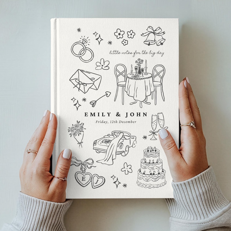May include: A white hardcover book with black line art illustrations of wedding icons including rings, a table setting, a car, and a cake. The text reads little notes for the big day, EMILY & JOHN, and Friday, 12th December, in a clean font.