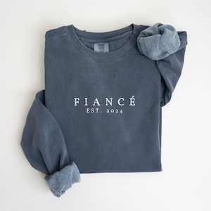 May include: A dark blue sweatshirt with the text "FIANC&Eacute; EST. 2024" in white.