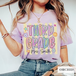 May include: A light purple t-shirt featuring a graphic with yellow vertical stripes and the text "THIRD GRADE" in a colorful, bubble-style font. Below the text, "Mrs. Smith" is written in black script, surrounded by small stars and flowers.