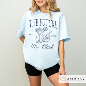 May include: Light blue t-shirt with a graphic of two cocktails and the text "The Future Est. 2024 Mrs. Clark" printed in navy blue.