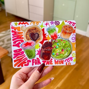 Könnte beinhalten: Eine rechteckige Karte mit einer farbenfrohen Illustration verschiedener Getränke, darunter Kaffee, Eiskaltgetränke und eine grüne Schale, mit dem Text FANCY DRINKS FOR THE WIN in kräftigen orangefarbenen und pinken Buchstaben und kleinen Sternen.