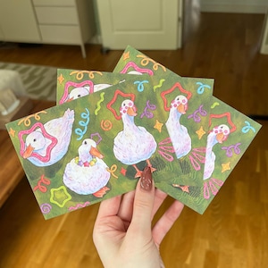 K&ouml;nnte beinhalten: Ein Set rechteckiger Karten mit einer wiederkehrenden Illustration von wei&szlig;en Enten mit orangefarbenen Schn&auml;beln und F&uuml;&szlig;en. Die Enten sind mit bunten sternf&ouml;rmigen Mustern verziert und von kleinen, lebhaften Kringeln und Sternen auf gr&uuml;nem Grund umgeben.