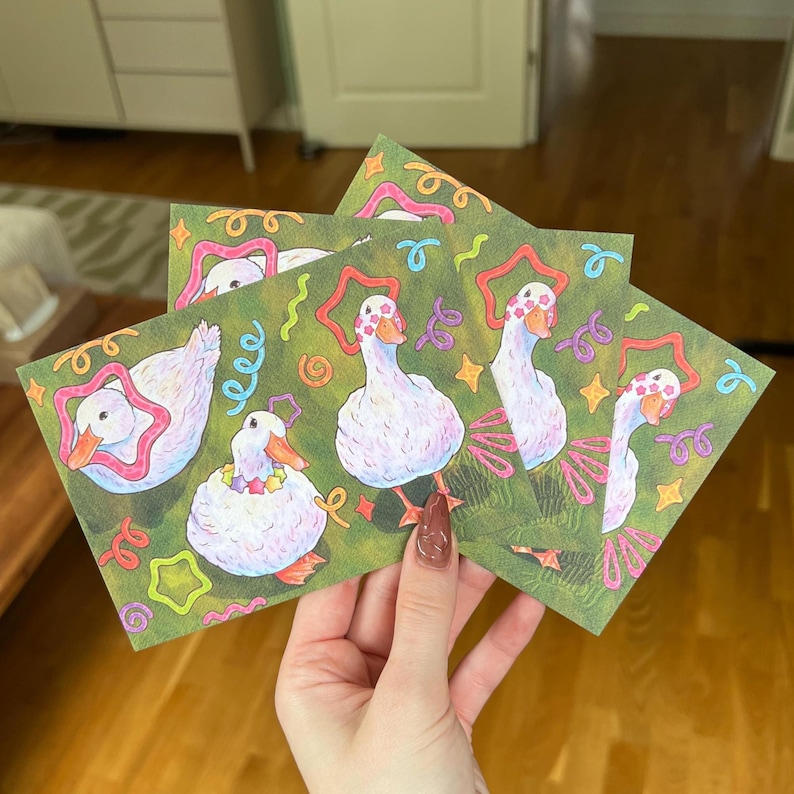 K&ouml;nnte beinhalten: Ein Set rechteckiger Karten mit einer wiederkehrenden Illustration von wei&szlig;en Enten mit orangefarbenen Schn&auml;beln und F&uuml;&szlig;en. Die Enten sind mit bunten sternf&ouml;rmigen Mustern verziert und von kleinen, lebhaften Kringeln und Sternen auf gr&uuml;nem Grund umgeben.
