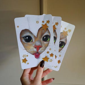 Könnte beinhalten: Drei weiße Karten mit einem Katzengesicht, das durch einen herzförmigen Ausschnitt blickt. Die Katze hat orangefarbenes Fell, grüne Augen und die Zunge ist herausgestreckt. Goldene Sterne sind um den Ausschnitt herum verstreut.