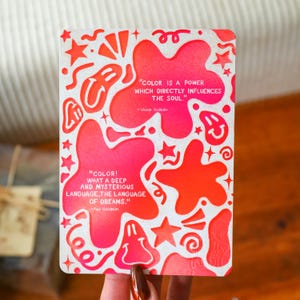 Könnte beinhalten: Eine rechteckige Karte mit weißem Hintergrund und einem Design aus roten und rosa Formen und Sternen. Die Karte enthält Zitate von Wassily Kandinsky und Paul Gauguin über Farbe und Träume.