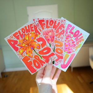 May include: Three rectangular greeting cards featuring vibrant illustrations of flowers in orange, pink, and red. Each card displays the text "A FLOWER FOR YOU" in bold, stylised lettering, accented with small star shapes scattered across the designs.