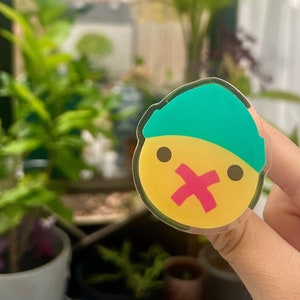 May include: A yellow sticker with a cartoon face wearing a teal beanie and a red X over its mouth. The sticker is clear and has a glossy finish.