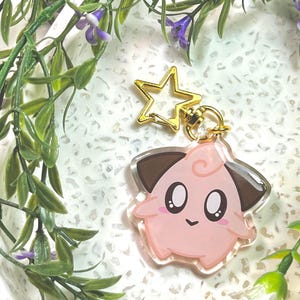 May include: A clear acrylic keychain featuring a cartoon character with a round pink body, dark brown ears, and large black eyes. The keychain is attached to a gold-colored metal star-shaped clasp, providing a secure way to attach it to items.