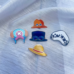 May include: Five colourful enamel pins featuring different hats from the anime One Piece. The pins are on a white fabric background.