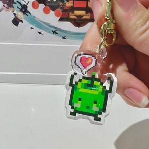 May include: A clear acrylic charm featuring a pixelated green creature with a heart-shaped thought bubble. The charm is attached to a gold-colored clasp. The background shows a colorful illustration.