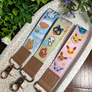 May include: Three colorful keychains with different cartoon characters. The keychains are made of fabric and have a brown leather strap with a gold metal clasp. The characters on the keychains are a bunny, a cat, and a fox.