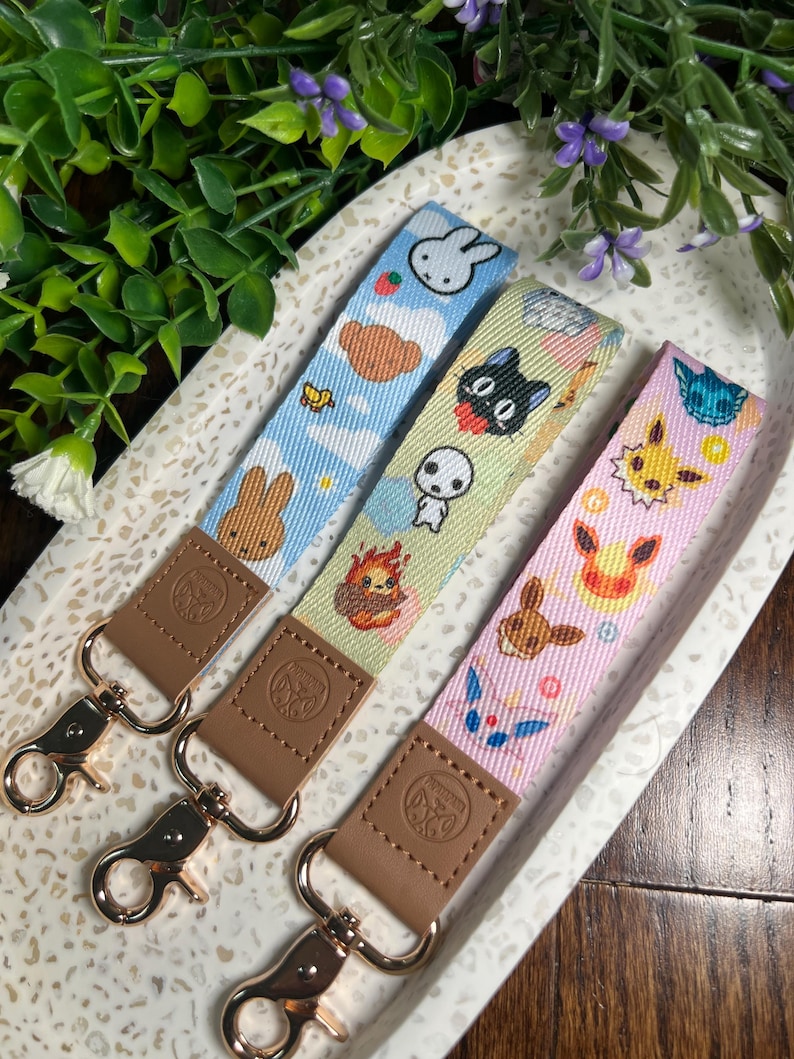 May include: Three colorful keychains with different cartoon characters. The keychains are made of fabric and have a brown leather strap with a gold metal clasp. The characters on the keychains are a bunny, a cat, and a fox.