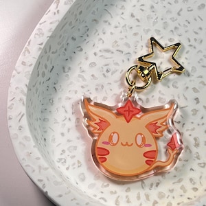 Puede incluir: Un llavero acrílico transparente con un diseño de criatura de dibujos animados en naranja y rojo, un cierre en forma de estrella dorada y una cadena dorada. El llavero está sobre una superficie blanca moteada.